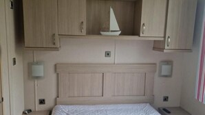 A hotel room with a bed, wooden cabinets, a nightstand, and a wall-mounted lamp. - Inviting 2-bed Caravan on Combe Haven Holiday Park (St Leonards-On-Sea)