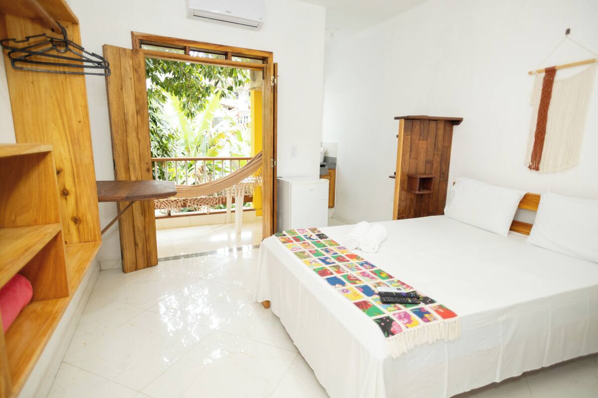 A hotel room with a bed, a wooden desk, a hammock, a wardrobe, and a view of greenery outside.