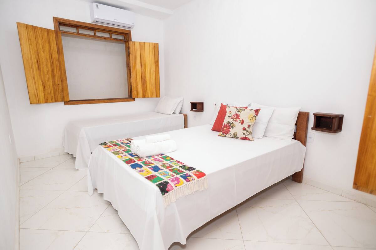 A hotel room with a bed, a smaller bed, a wooden-framed window, and a wall-mounted air conditioner.