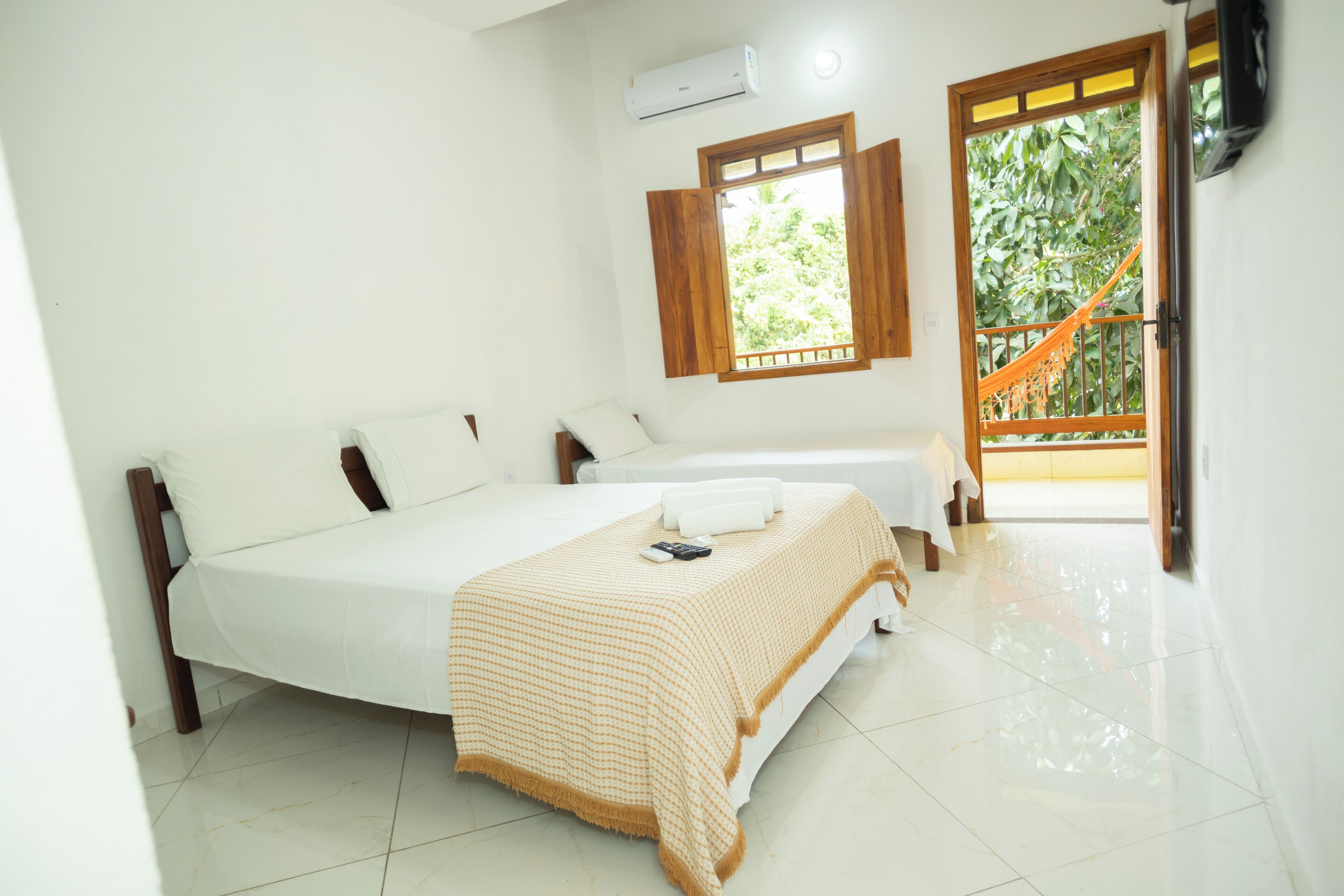 A bedroom with a bed, two pillows, a bedside table, a window with wooden shutters, and a view of greenery outside.