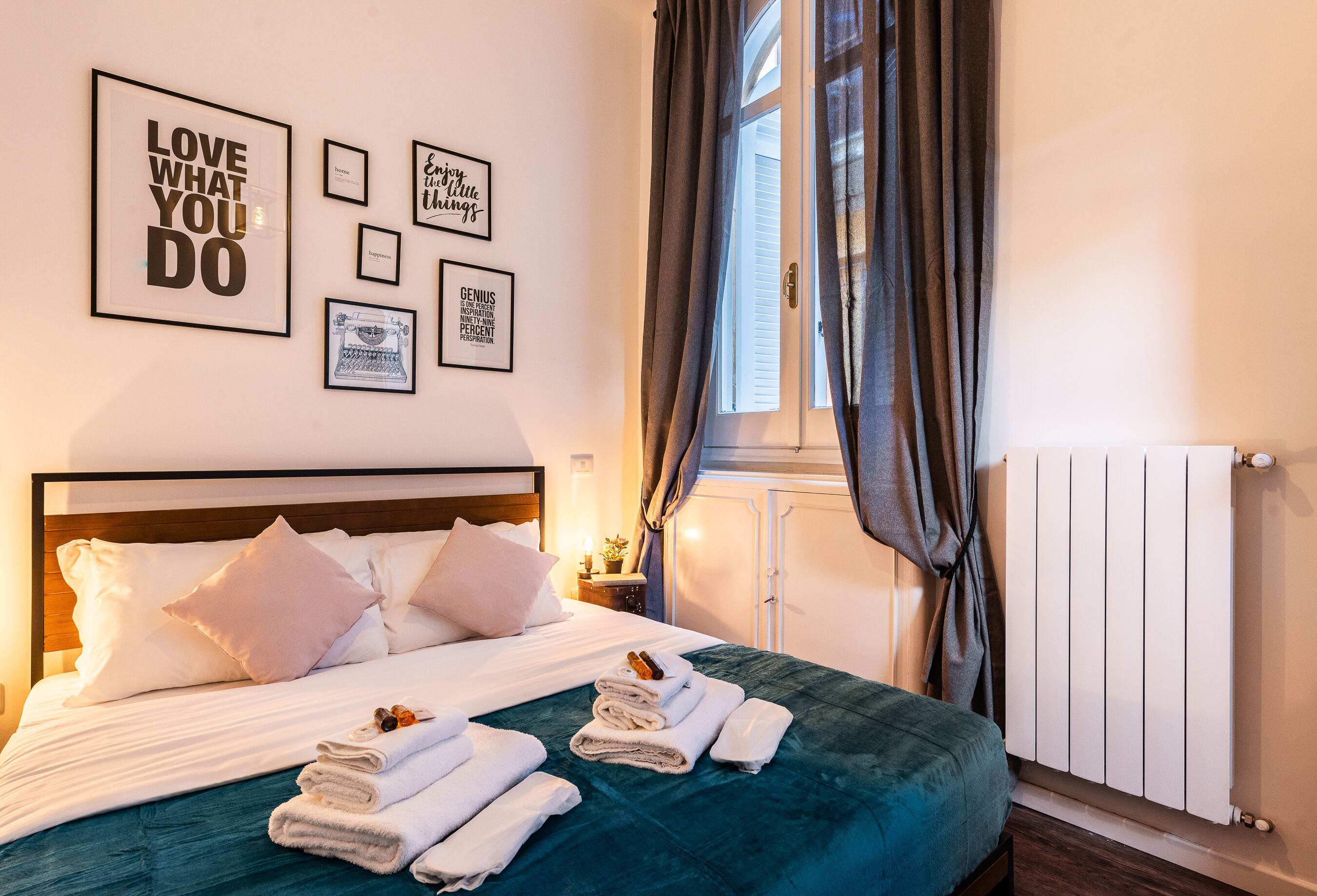 Een netjes opgemaakt bed met witte lakens en blauwgroene beddengoed, een muur met motiverende posters, een raam met gordijnen en een radiator.