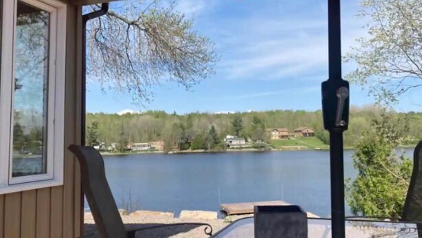 A lakeside patio with a red umbrella, outdoor seating, and a view of a lake and distant houses. - Spacious Waterfront in the French River

 (Noëlville)