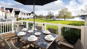 A deck with a table set for four, overlooking a river and houses. - Isis Lakes 50, Natterjack Lodge - Lakeside Lodge, On-Site Activities, Sleeps 6 (SOUTH CERNEY)