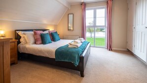 A bedroom with a large bed, a wooden nightstand, a mirror, and a view of a green landscape through a window. - Isis Lakes 50, Natterjack Lodge - Lakeside Lodge, On-Site Activities, Sleeps 6 (SOUTH CERNEY)
