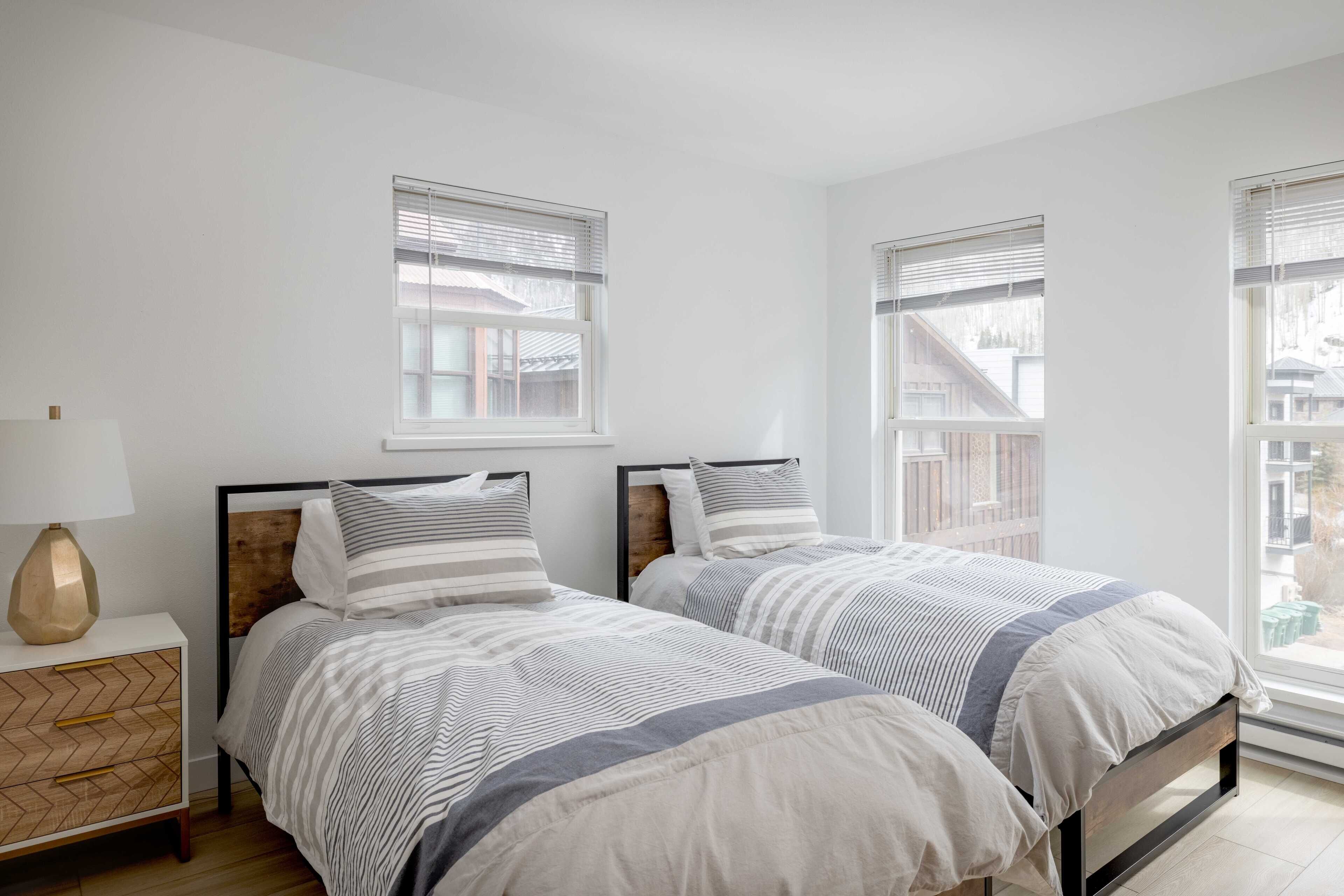 A bedroom with two beds, a nightstand with a lamp, and large windows with blinds.