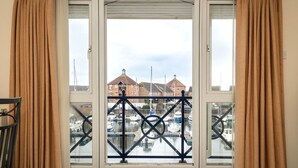 Une fenêtre avec des rideaux de couleur beige, une rambarde en métal et une vue sur une marina, avec des bateaux et des bâtiments en arrière-plan.