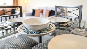 Une table à manger dressée avec un saladier blanc, des assiettes et des verres, agrémentée de coussins à motifs et d’une chaise au design moderne.