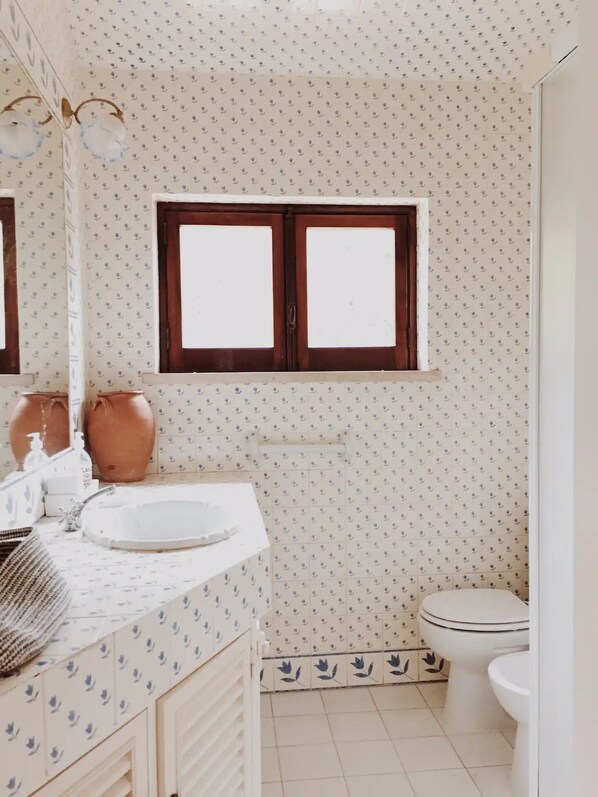 Un baño con lavamanos blanco, espejo, ventana y retrete.