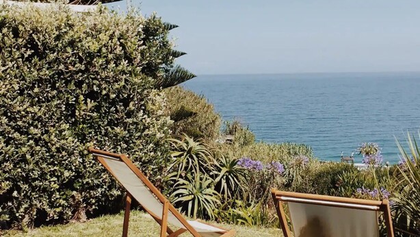 A beachside view with two wooden lounge chairs facing the ocean, surrounded by lush greenery and a clear blue sky. - Casa da Praia (Colares)