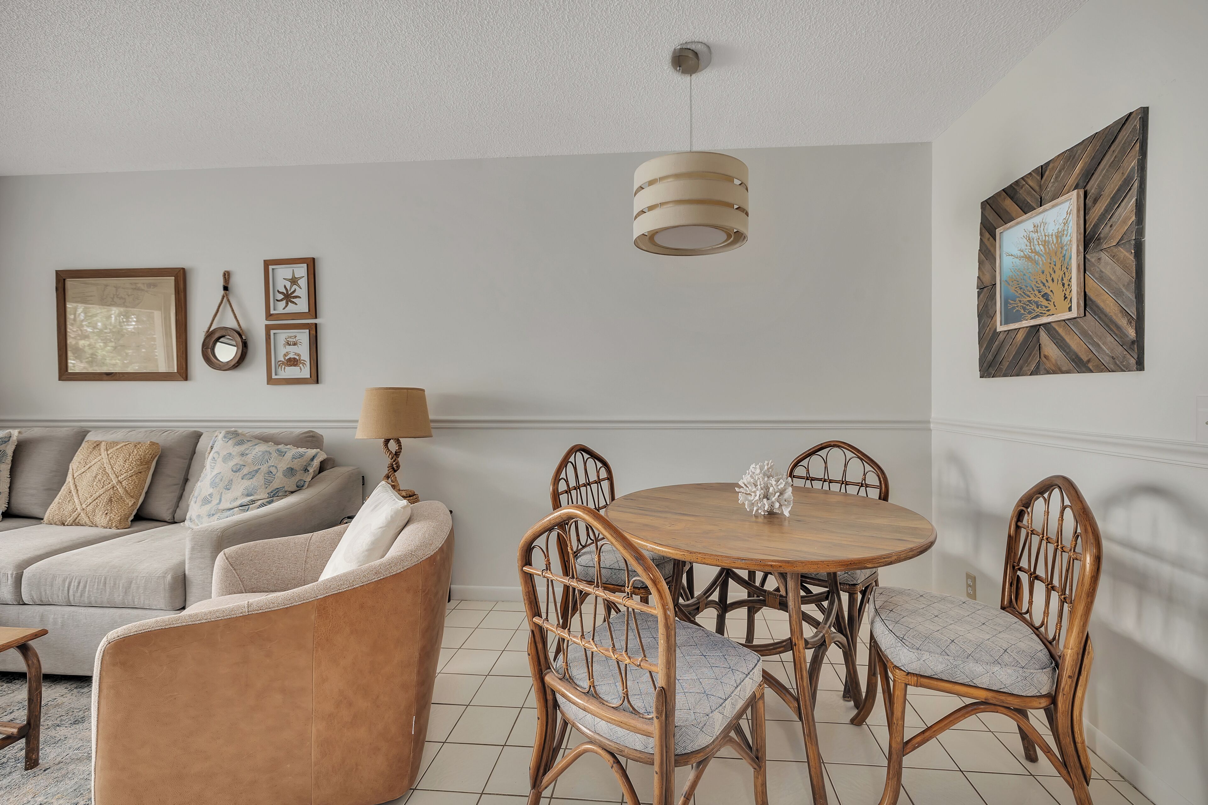 A dining area with a round wooden table and wicker chairs, a sofa with patterned cushions, and wall-mounted artwork.
