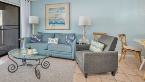 Una sala de estar con un sofá azul, un sillón gris, una mesa de centro de vidrio y dos lámparas.