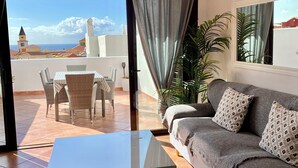 Una sala de estar con un sofá gris, cojines estampados, una mesa de centro de vidrio y vistas a una terraza con un comedor y una planta.