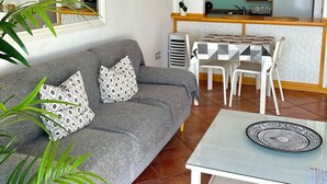 Una sala de estar con un sofá gris, cojines estampados, una mesa de centro de vidrio y una zona de comedor con mantel a cuadros.