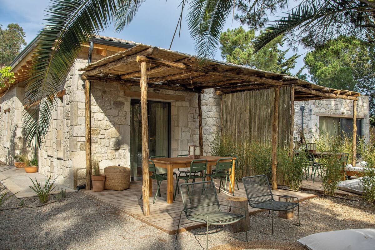 Ein rustikales Steinhaus mit einer Holzpergola, einem Tisch mit Stühlen und einem kleinen Garten mit Blumentöpfen.