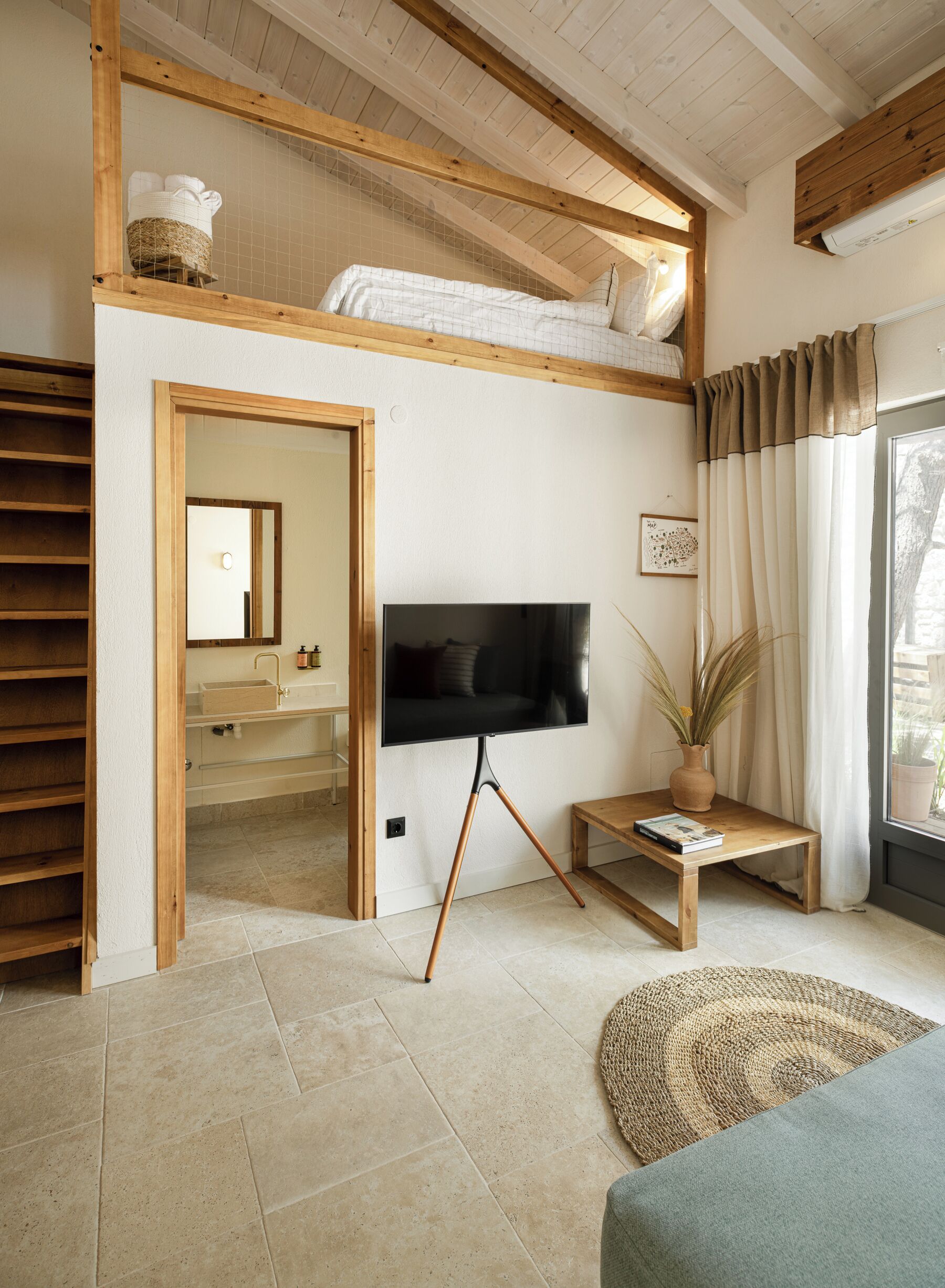 Un dormitorio tipo loft con un televisor sobre un soporte de trípode, un banco de madera, una alfombra tejida y vistas al exterior a través de una ventana.