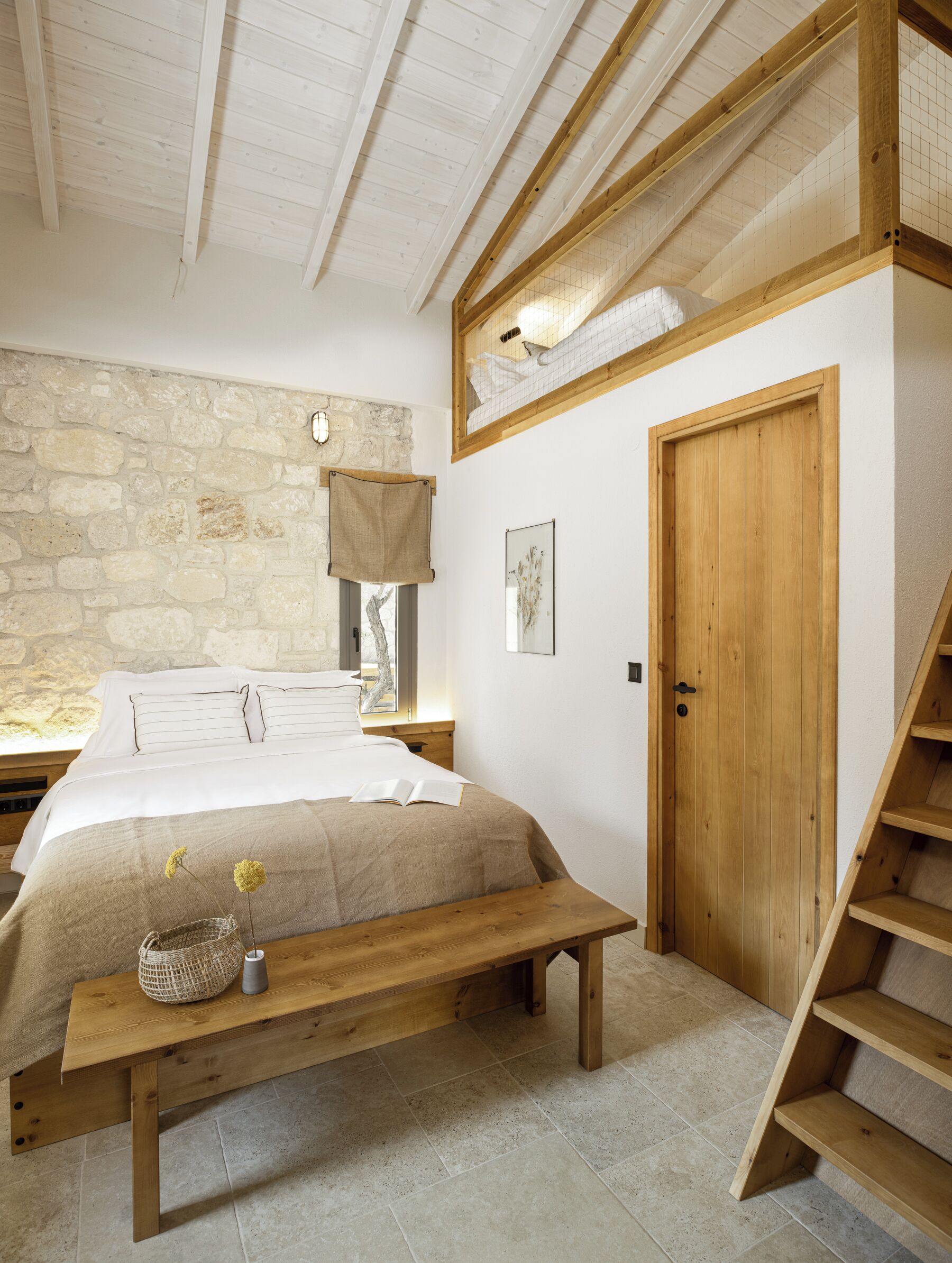 Une chambre avec un mur en pierre, un lit en bois, un banc, une porte en bois et un escalier.