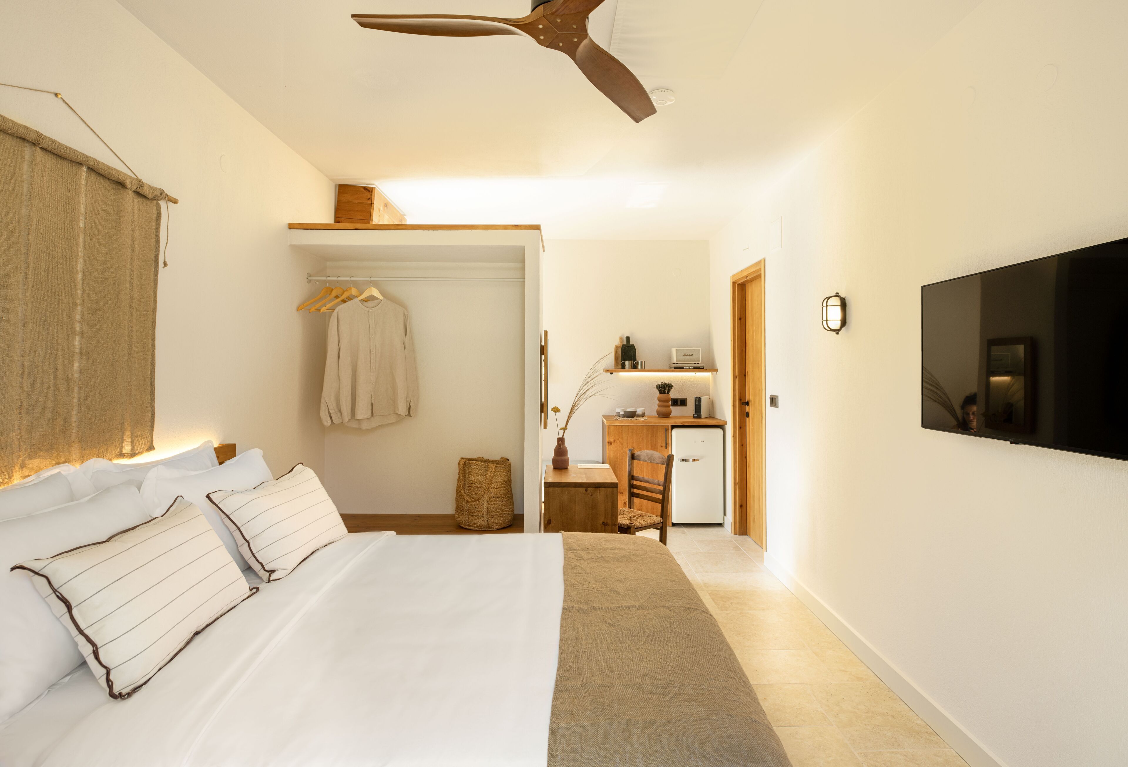 Un dormitorio con cama, ventilador de techo, televisor montado en la pared, escritorio con silla y una pequeña área de kitchenette.