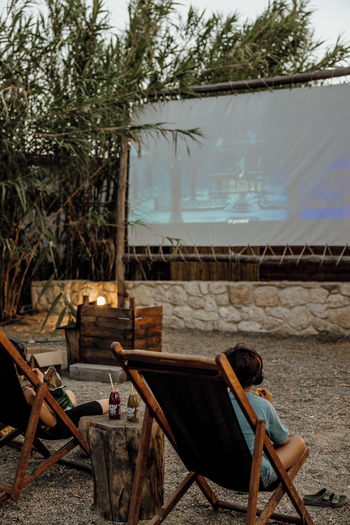 Two people seated on wooden chairs outdoors, watching a movie on a large screen.