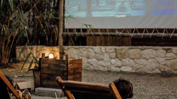 Two people seated on wooden chairs outdoors, watching a movie on a large screen.