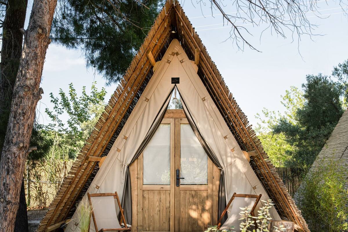 Eine dreieckige Holzhütte mit einer großen, zentralen Tür und zwei Fenstern, umgeben von Bäumen und einer Holzdielenveranda.