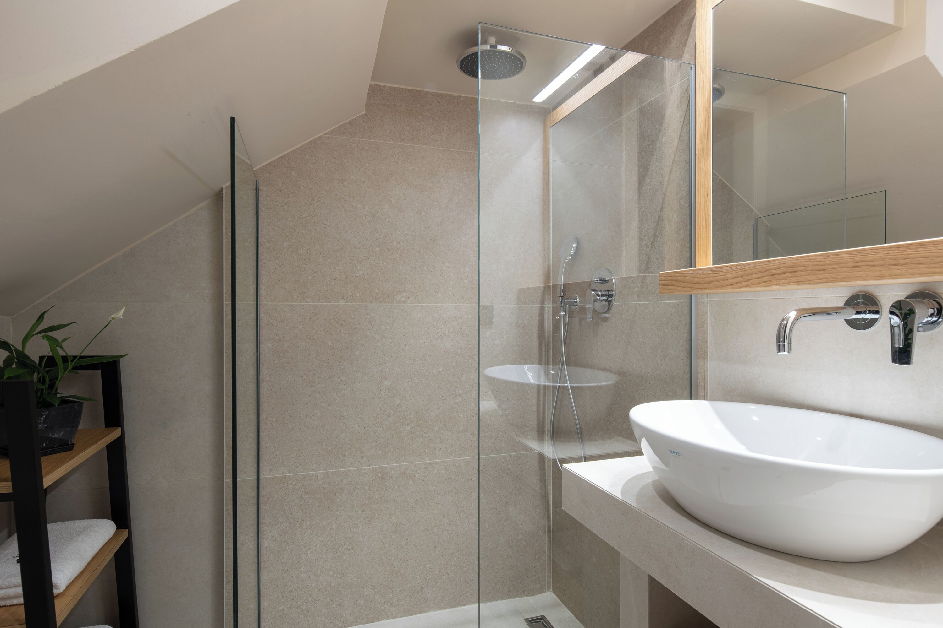 Un baño moderno con cabina de ducha de vidrio, un lavabo blanco y un estante de madera con una planta en maceta.