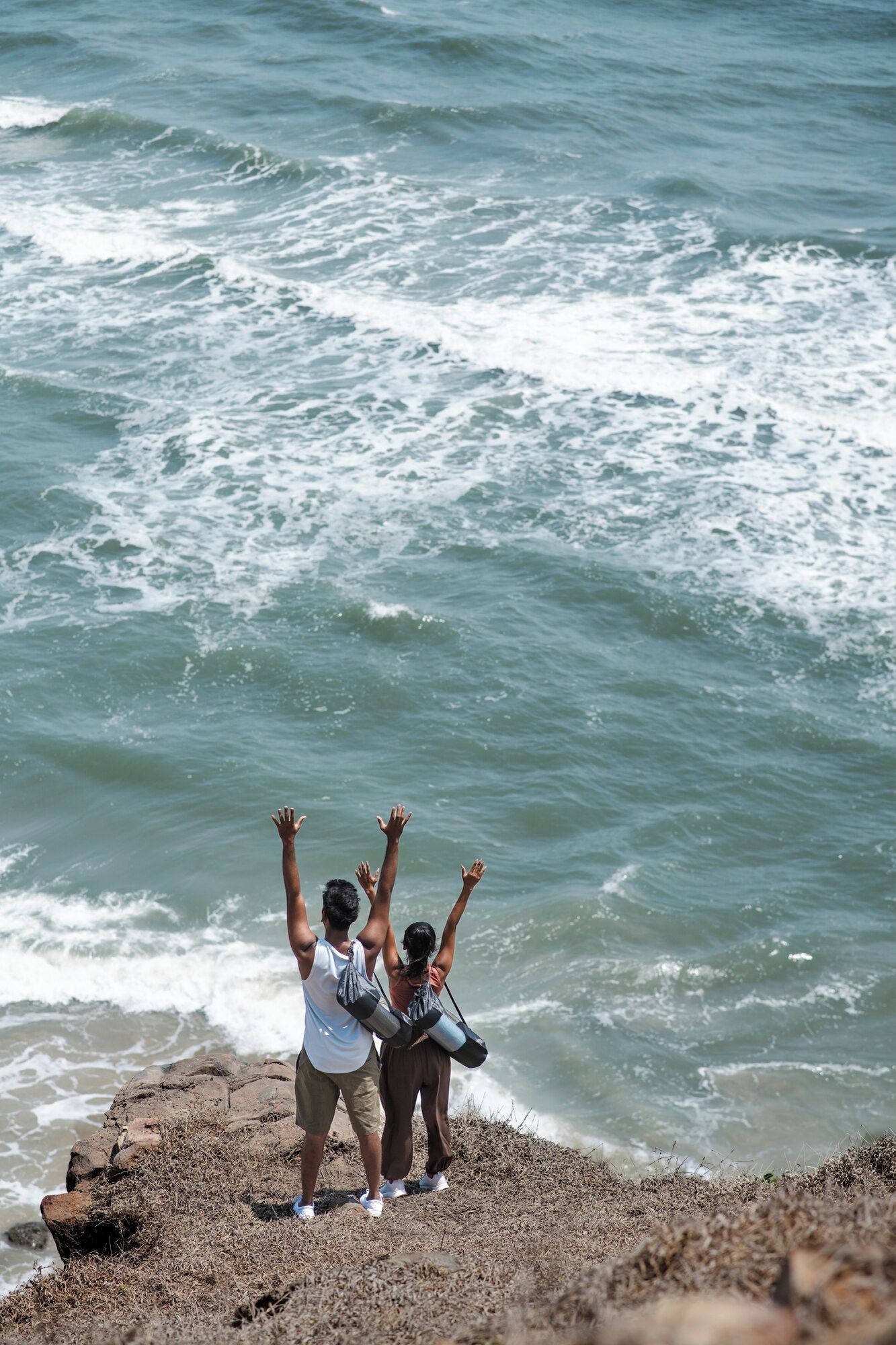Two people standing on a rocky shore with their arms raised, overlooking the ocean.