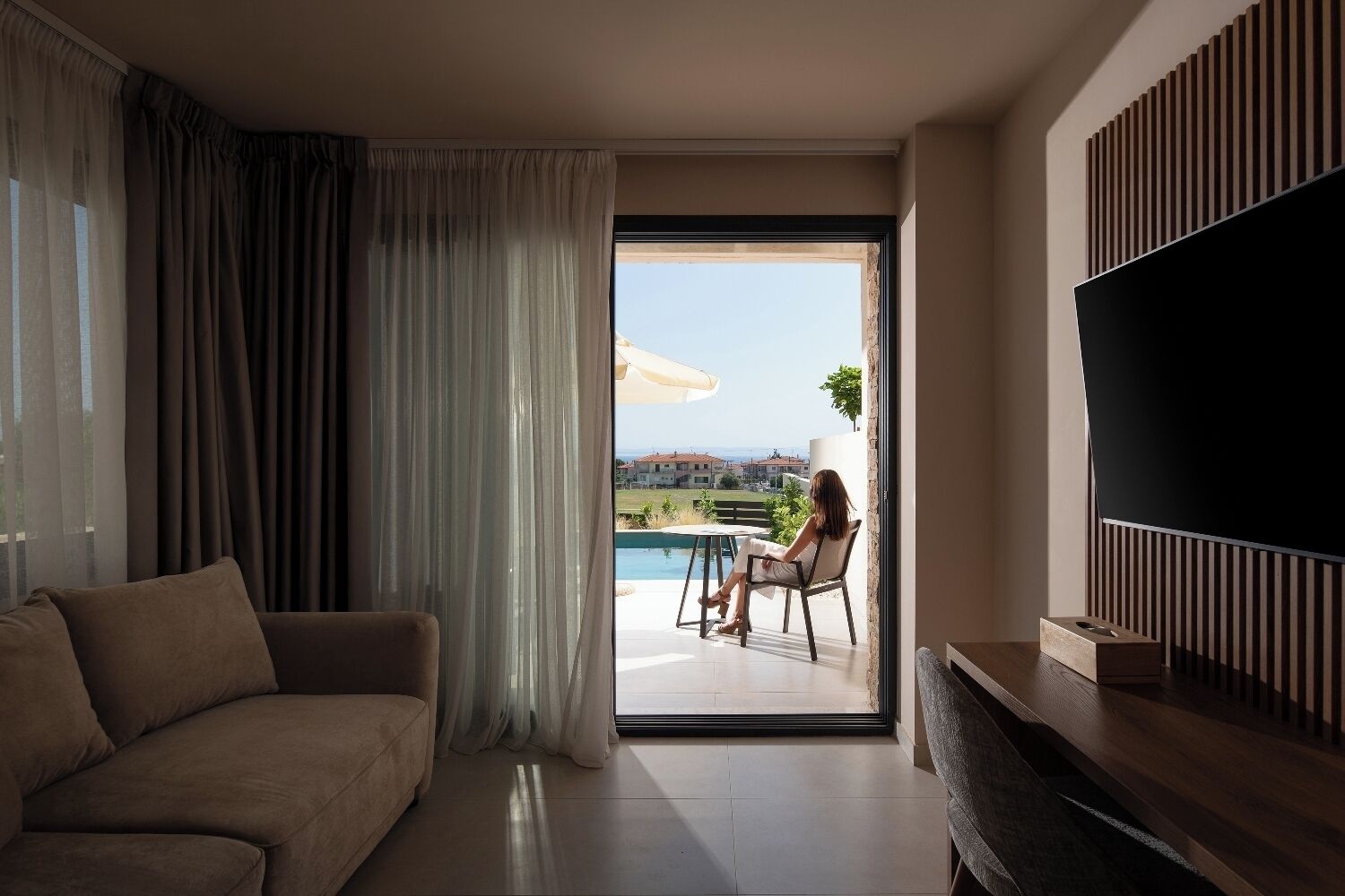 A modern living room with a sofa, a TV, and a sliding glass door leading to a balcony with a pool.