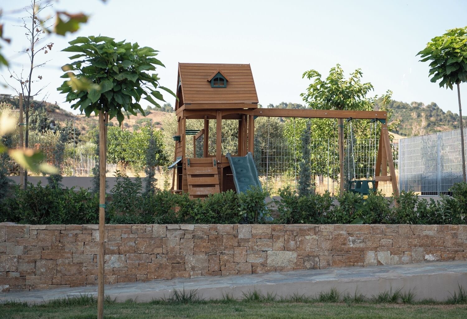 A wooden play structure with a slide and climbing area, surrounded by a stone wall and greenery.