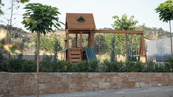 A wooden play structure with a slide and climbing area, surrounded by a stone wall and greenery.