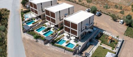 A modern multi-story building with three separate swimming pools, surrounded by a landscaped garden and a gravel driveway.