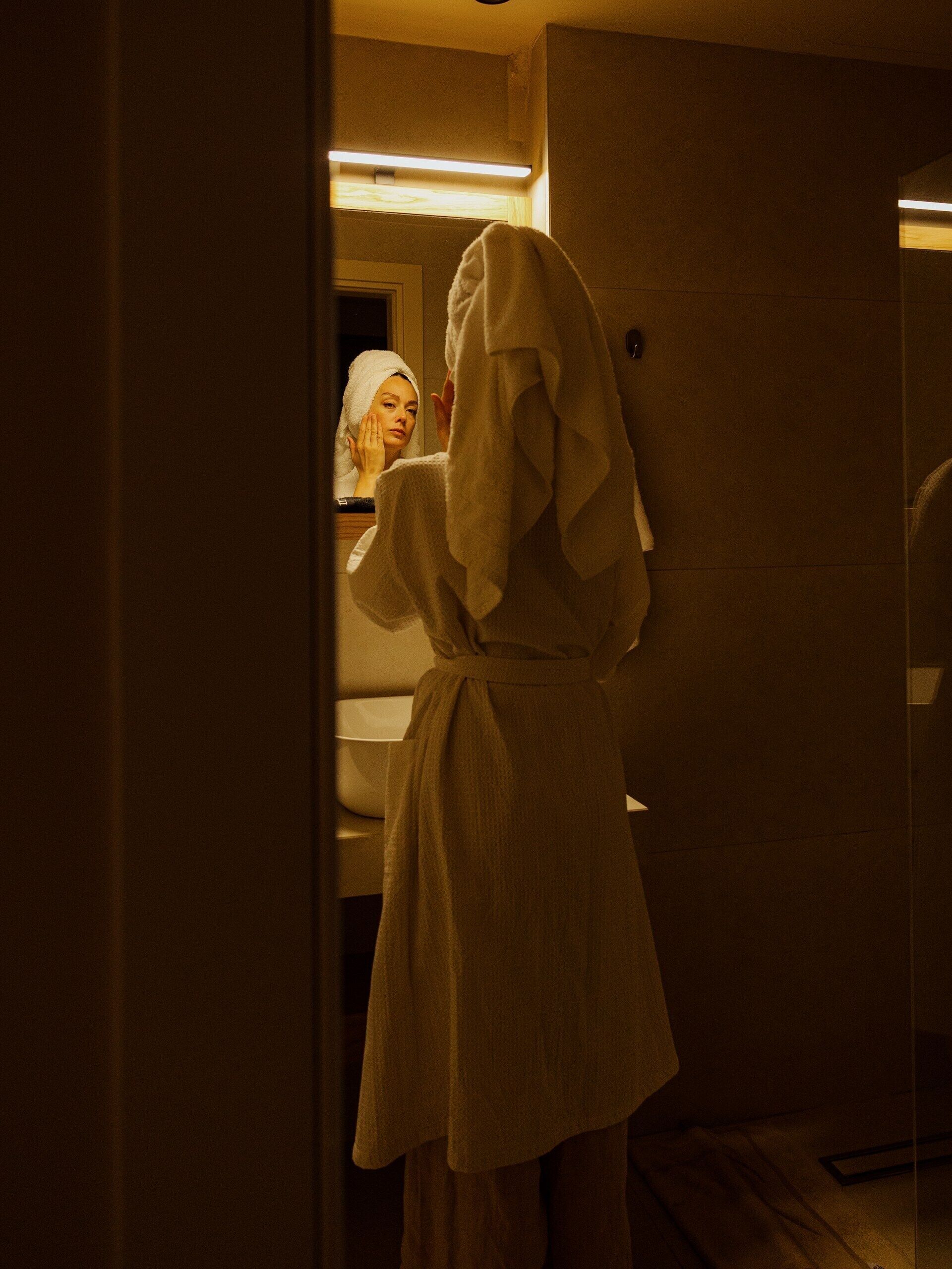 A person in a white robe standing in a bathroom, looking into a mirror.