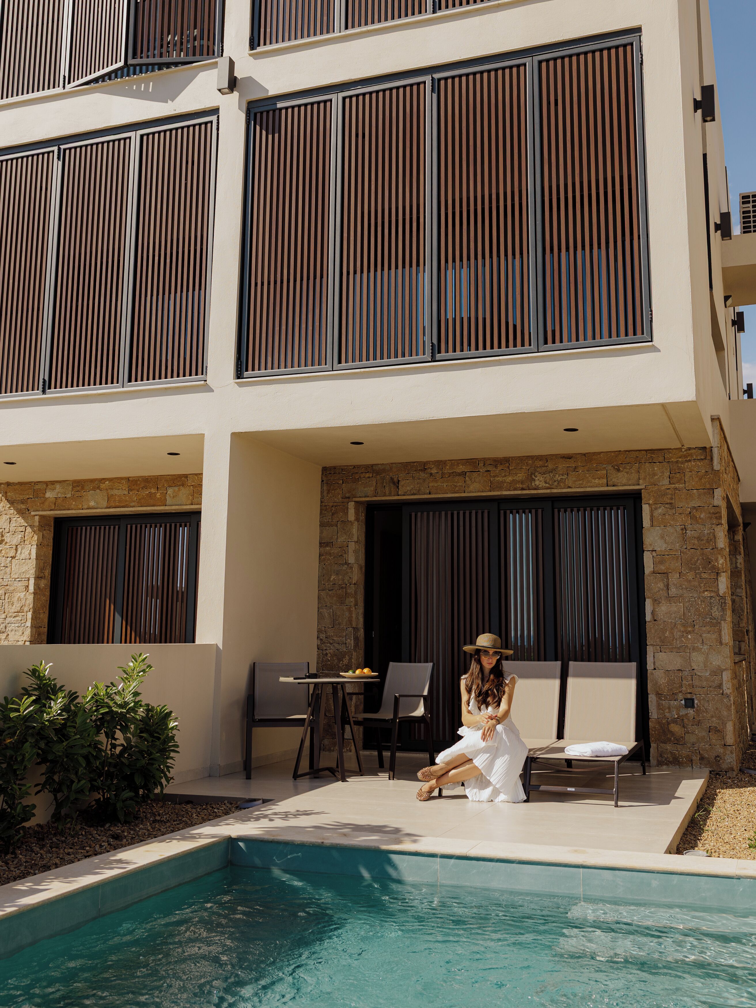 A modern building with a pool area, featuring a seating area with chairs and a table, and a person sitting by the pool.