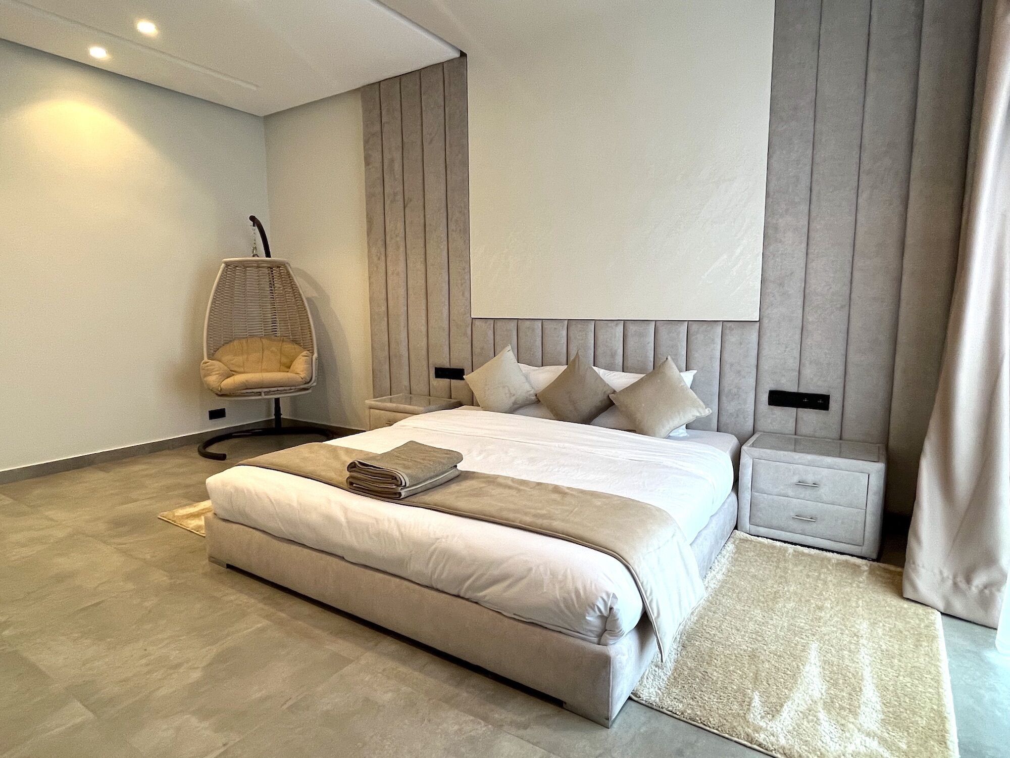 A modern bedroom with a large bed, a hanging chair, and a nightstand.