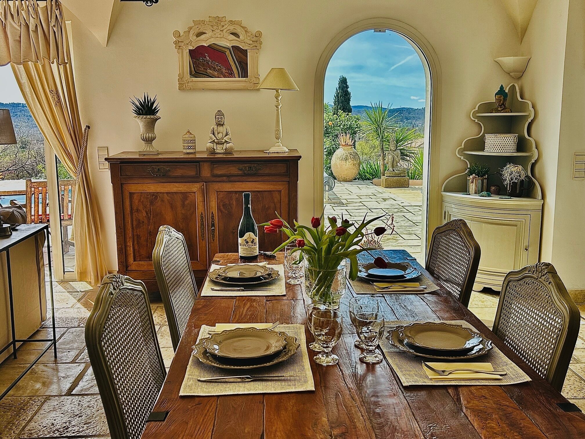 A dining area with a wooden table set for four, a Buddha statue, and a view of a garden through an arched doorway.