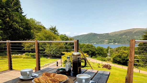 Uma mesa de madeira com um prato de panquecas, duas xícaras de café e uma cafeteira numa varanda com vista para um lago e colinas.