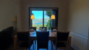 Uma área de refeições com uma mesa posta para dois, duas cadeiras, uma janela com vista para vegetação e colinas, e duas luminárias de mesa.