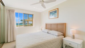 A bedroom with a bed, a nightstand, a lamp, a ceiling fan, a window with a view of the beach, and a framed picture on the wall. - Pacific Gables 1 - Absolute Beachfront Tugun - Linen Provided - Min. 3 Night Stays! (Tugun)