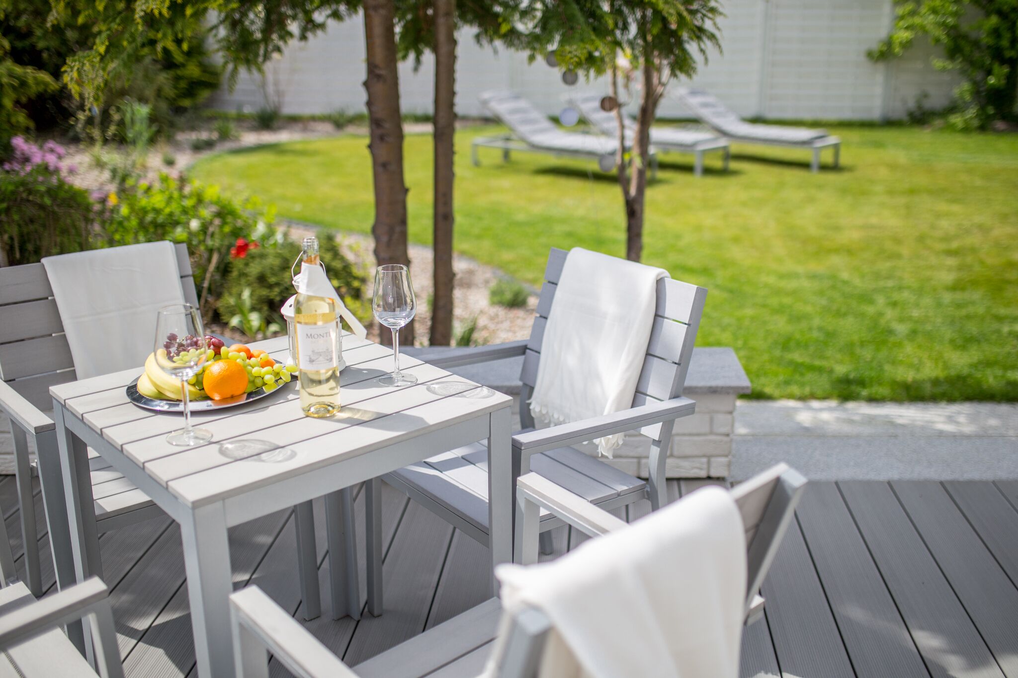A patio table with a fruit platter, wine bottle, and glasses, surrounded by white chairs.