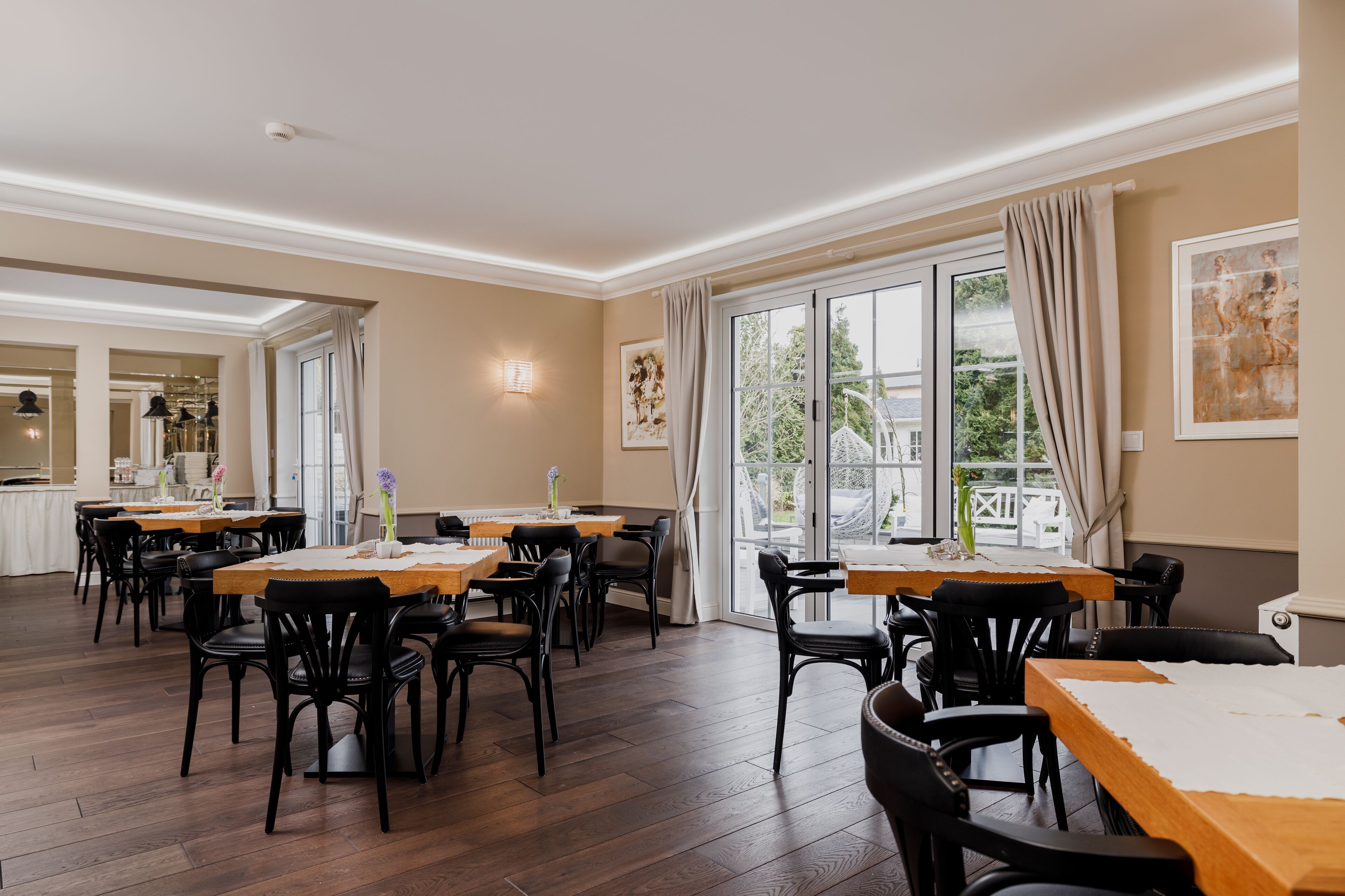 A dining area with multiple tables and chairs, large windows with curtains, and artwork on the walls.