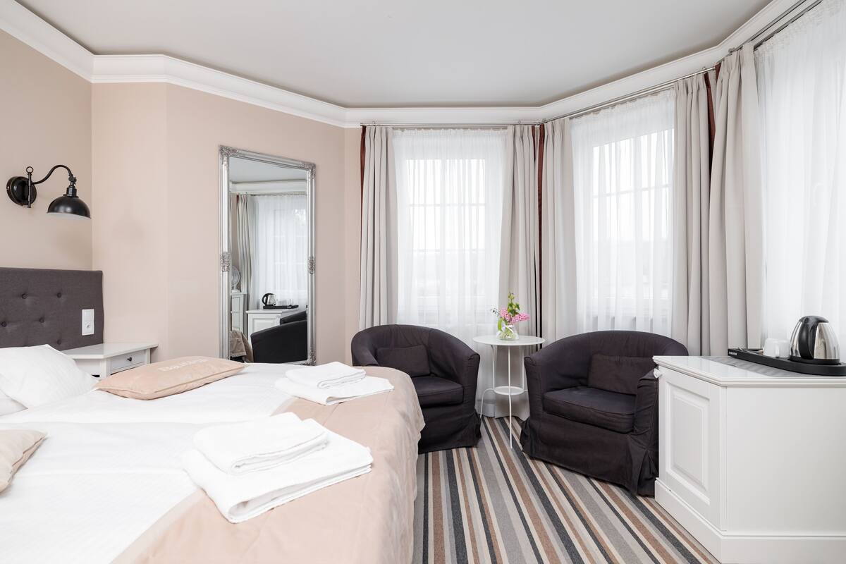 Habitación de hotel con una cama grande, dos sillones, una mesita con un jarrón de flores y un ventanal con cortinas.