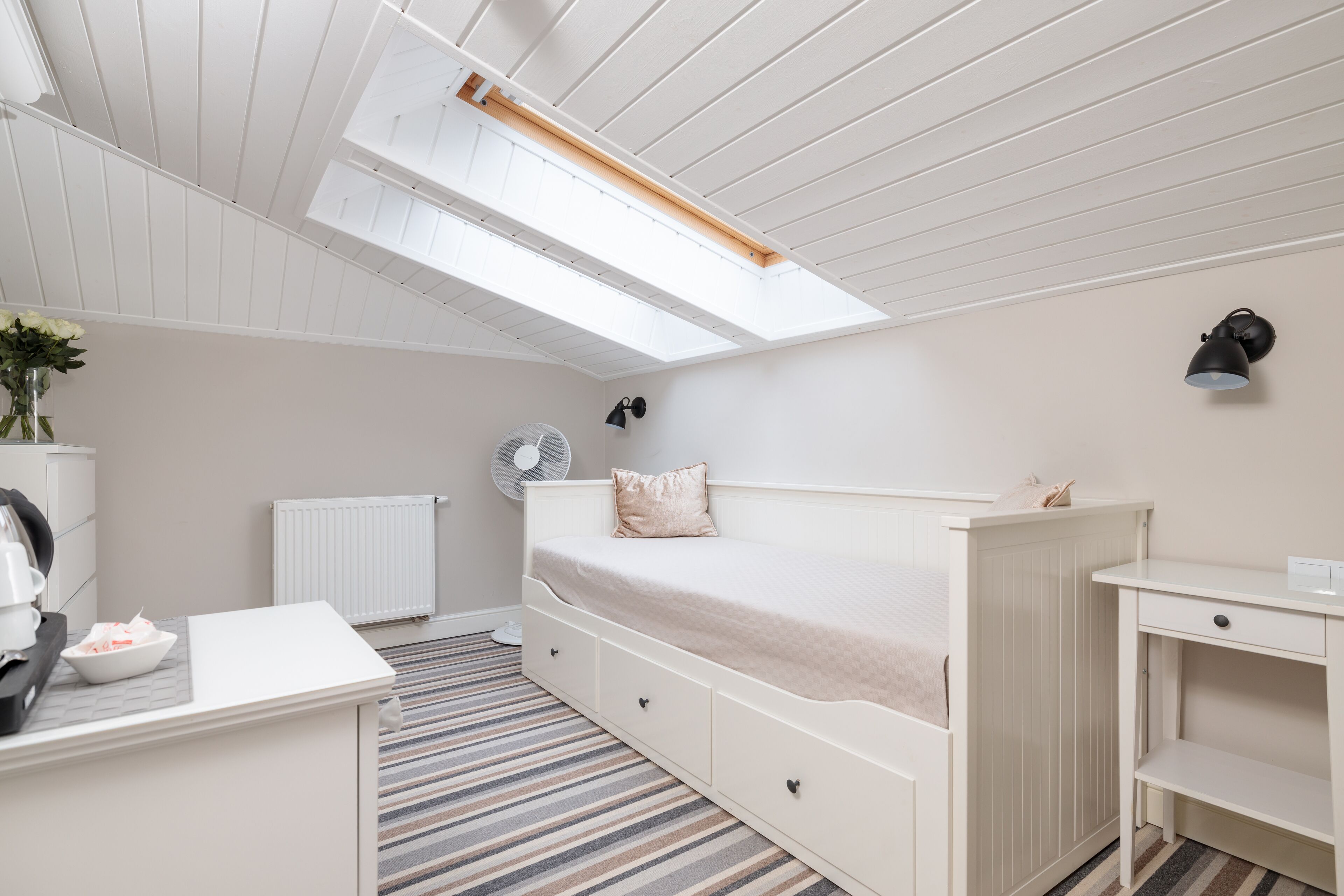 Un dormitorio con una cama, un escritorio, un radiador, un ventilador y un tragaluz.