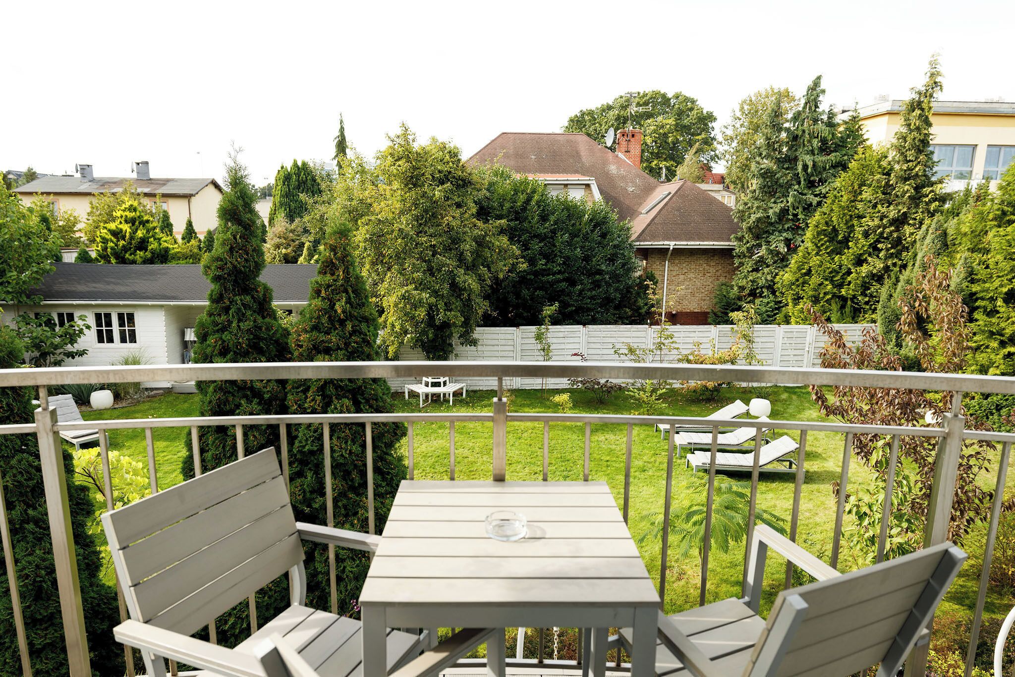 A balcony with a table and two chairs, overlooking a well-maintained garden and neighboring houses.