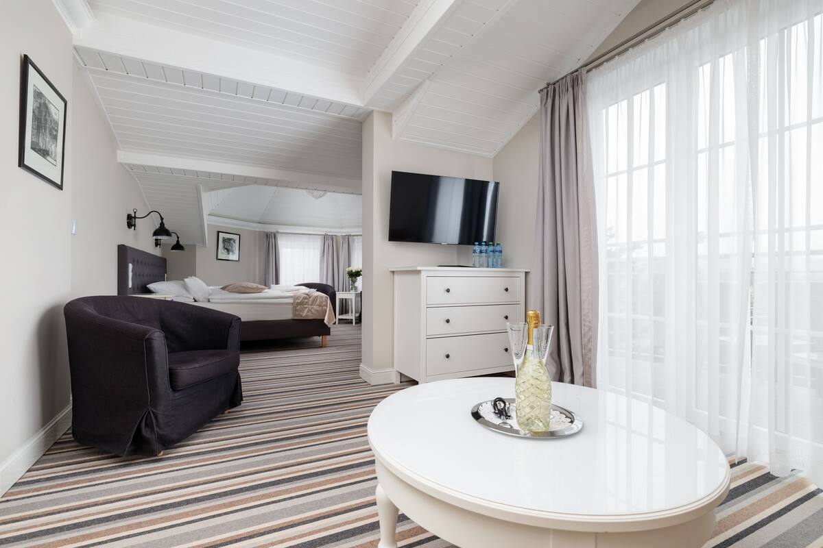 Habitación de hotel con cama, sofá, televisor, mesa de comedor con bandeja y un ventanal con cortinas.