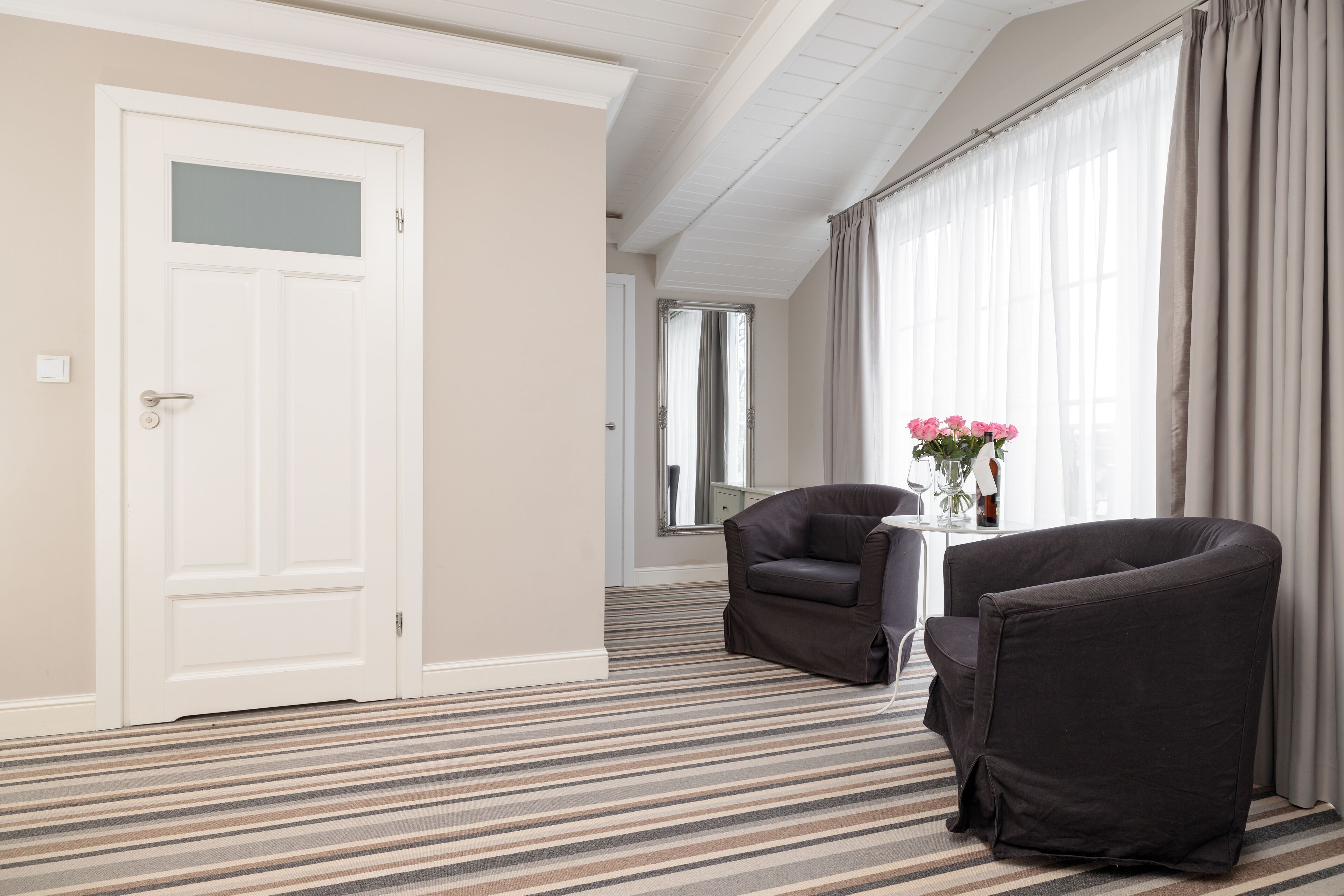 A hotel room with a striped carpet, a white door, a dark armchair, a small table with a vase of flowers, and a large window with curtains.