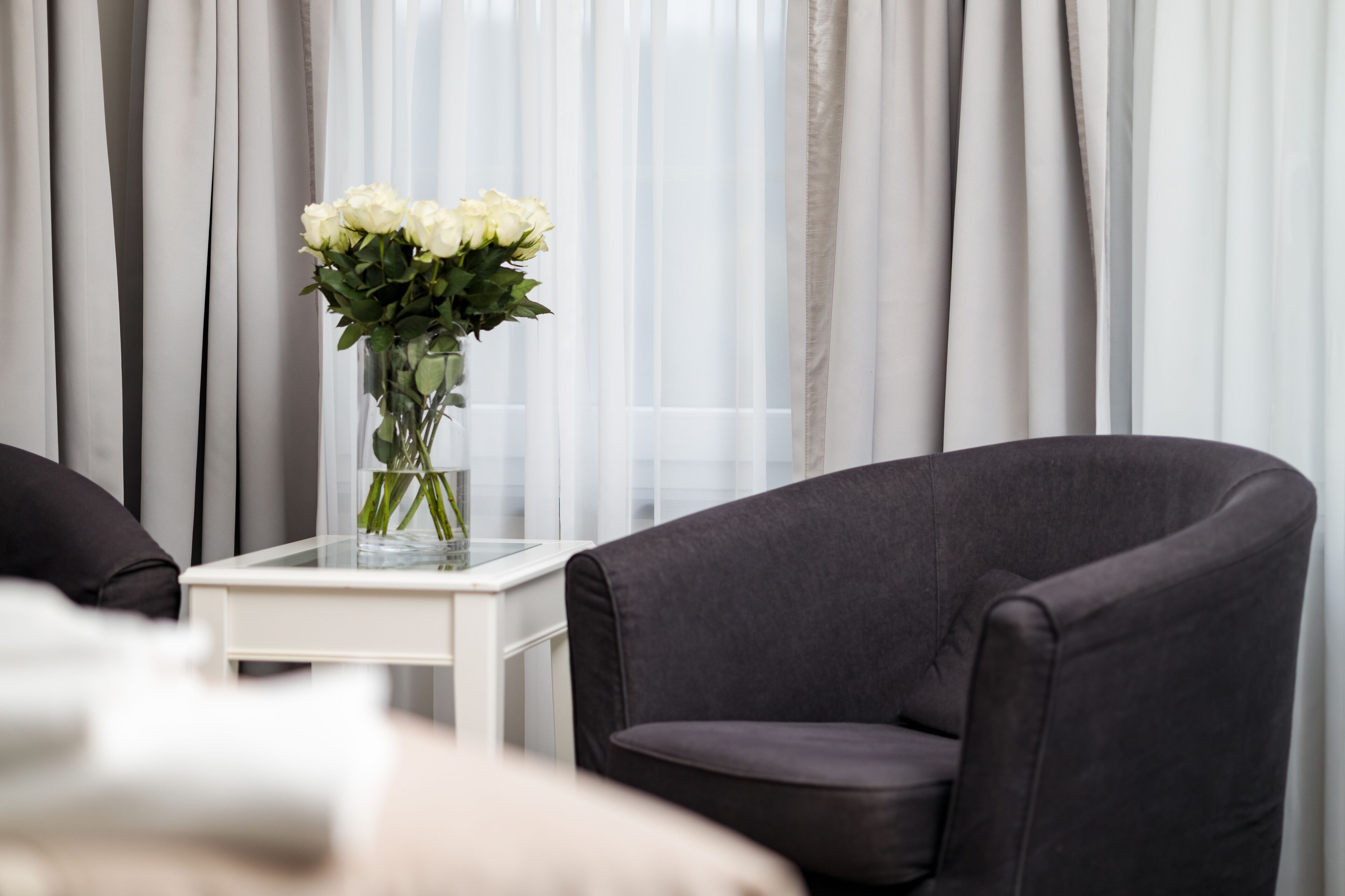 A modern living room with a dark grey armchair, a glass coffee table with a vase of white flowers, and sheer white curtains in the background.