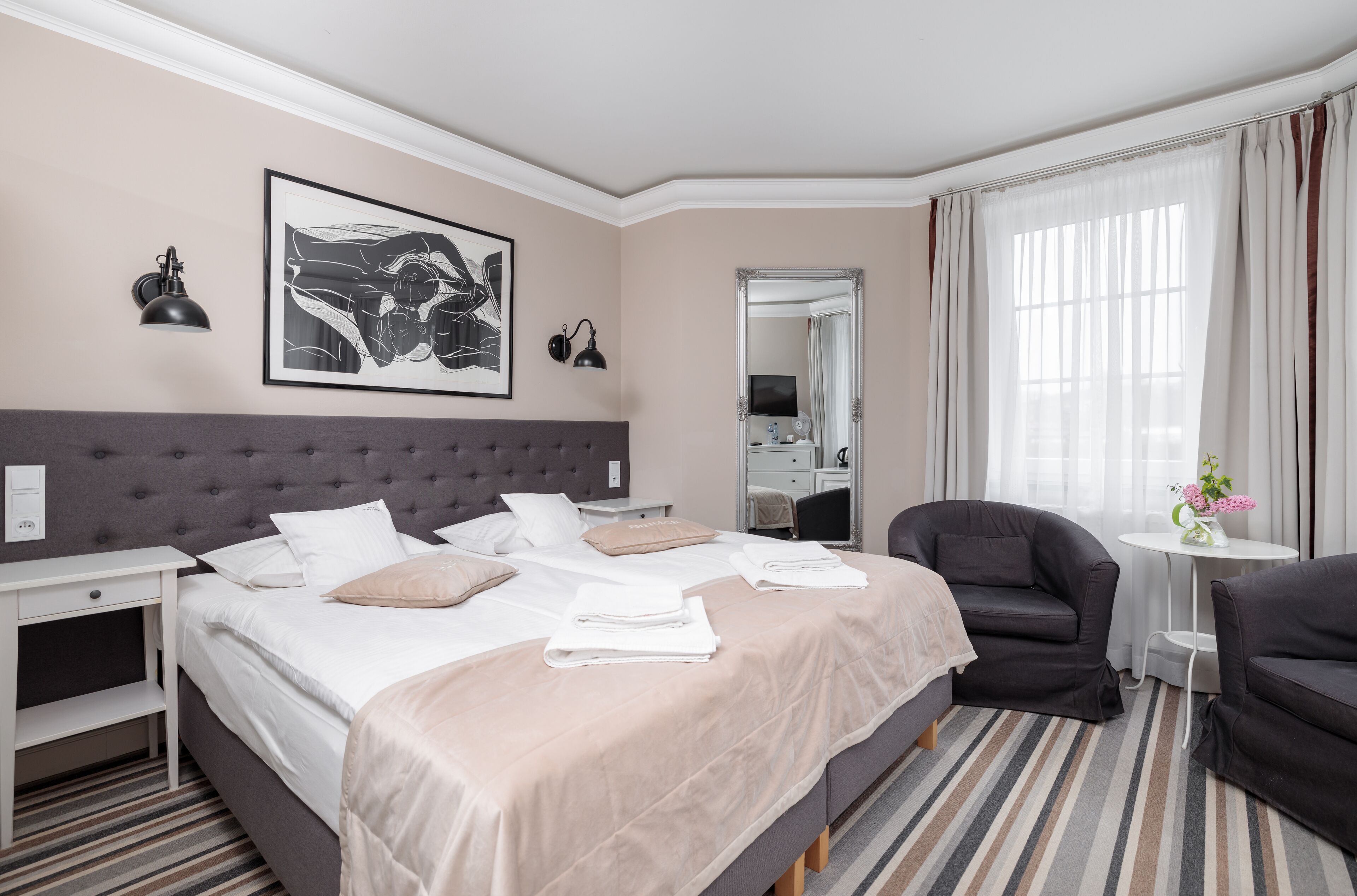 Habitación de hotel con una cama grande, dos mesitas de noche, una silla, una pequeña mesa redonda con un florero y un espejo, además de una obra de arte en la pared.