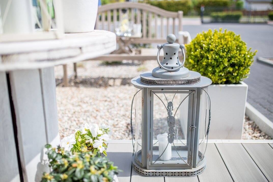 A clear glass lantern with a white base, a decorative top, and a small figure inside, placed on a wooden deck.
