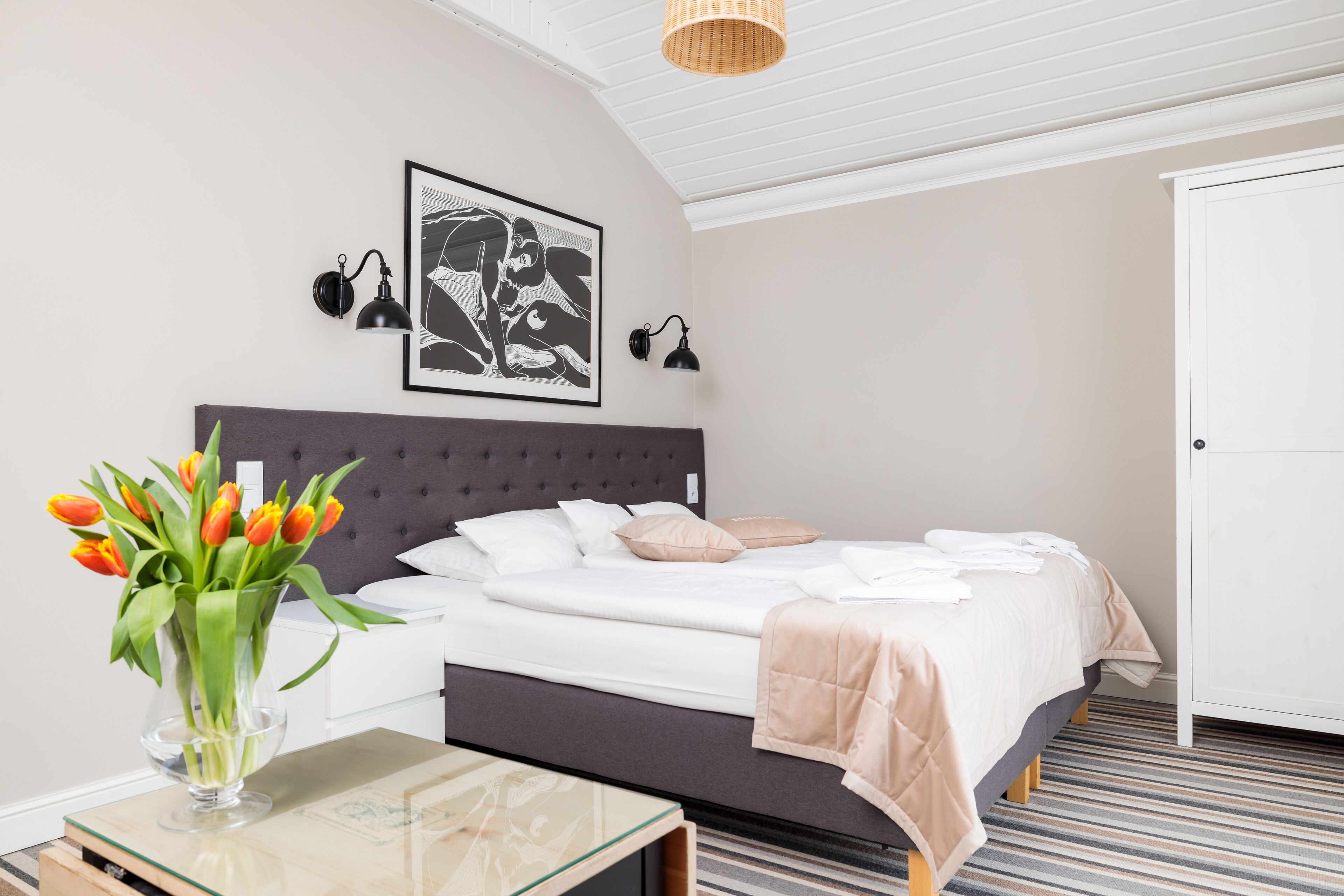 A modern bedroom with a tufted headboard, a bed with white linens, a vase of tulips, and a striped carpet.