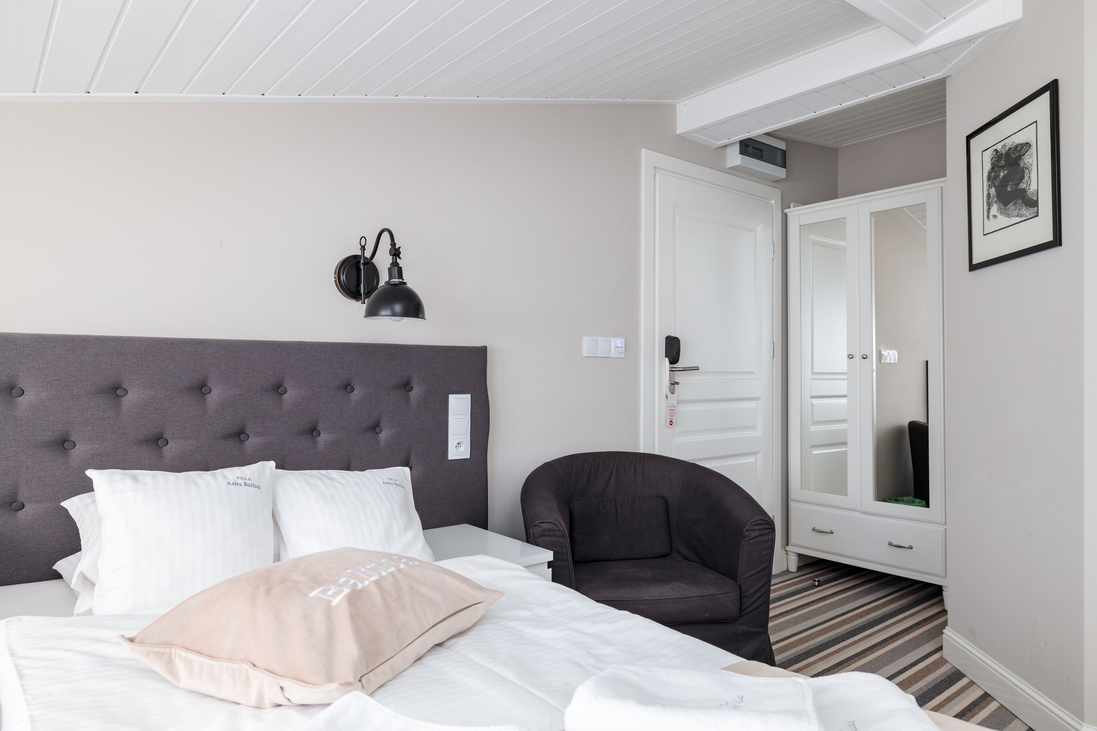Ett hotellrum med en stoppad sänggavel, en säng med vita och beige kuddar, en mörk fåtölj, ett vitt sängbord, ett spegelskåp och ett inramat konstverk på väggen.