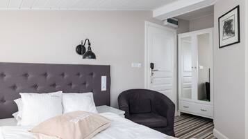 Ett hotellrum med en stoppad sänggavel, en säng med vita och beige kuddar, en mörk fåtölj, ett vitt sängbord, ett spegelskåp och ett inramat konstverk på väggen.